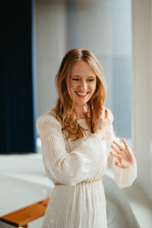 Woman smiling in sunlit room, elegant white dress.