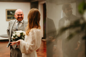 Bride holding bouquet facing groom, reflection, warm wedding ambiance.