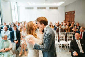 Couple kissing at wedding ceremony, guests watching.