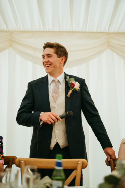 Groom smiling during wedding speech in marquee.
