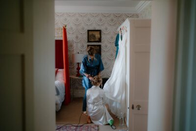 Bride getting ready in bedroom, dress hanging nearby.