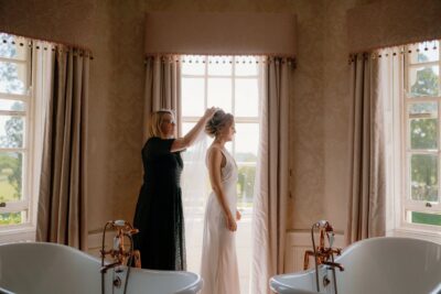 Woman adjusts bride's veil near window, elegant bathroom.