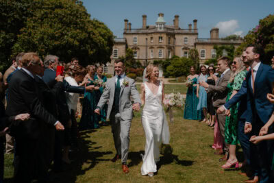 Newlyweds walk through guests outside stately home.