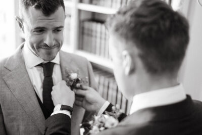 Groomsman pinning boutonniere on smiling groom's suit.