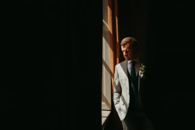 Groom in sunlight by window, thoughtful expression.