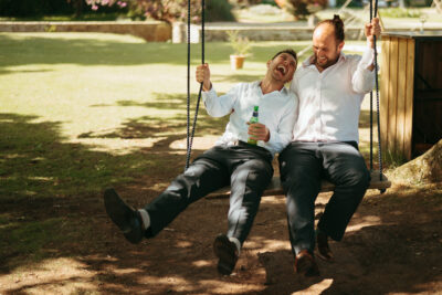 Two men laughing on swing with drinks in garden.