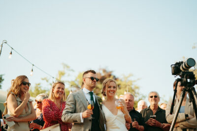 Outdoor wedding toast with smiling guests and camera.