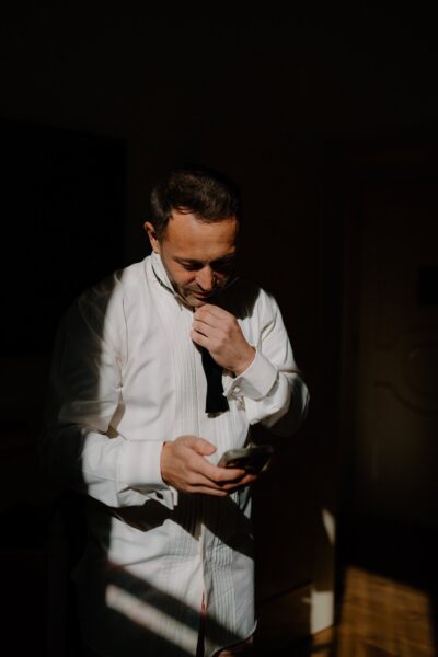 Man adjusting tie in natural light, holding phone.