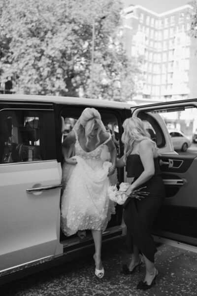 Bride exits car assisted by bridesmaid.