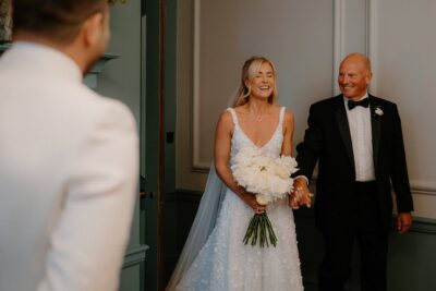 Bride holding father's hand, smiling in hallway.