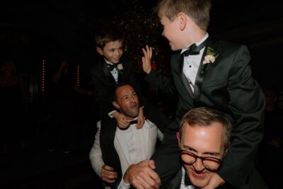 Two boys in suits on men’s shoulders at event.