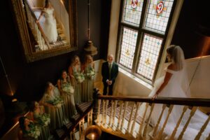 Bride on stairs with bridesmaids and stained glass window.