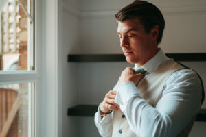 Man adjusting tie by window, formal attire.