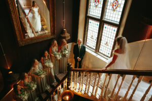 Bride descending staircase with bridesmaids and father watching.
