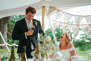 Groom giving speech, bride laughing at wedding reception.