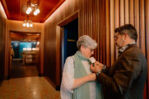 Man adjusts corsage on woman's outfit indoors.