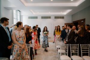 Woman walks down aisle at indoor wedding ceremony.
