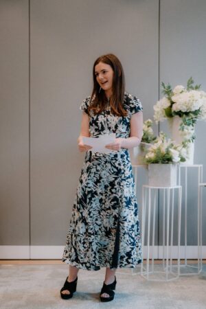 Woman speaking at event with flowers in background.