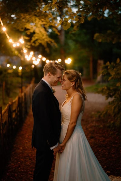 Bride and groom smiling under fairy lights in woods.