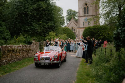 Red wedding car departs church, crowd cheers.