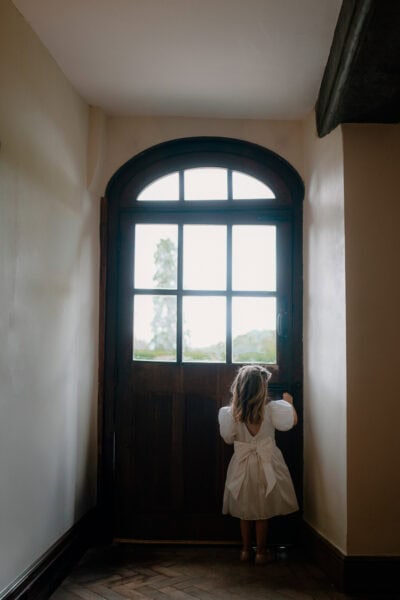 Child in white dress looks out wooden door