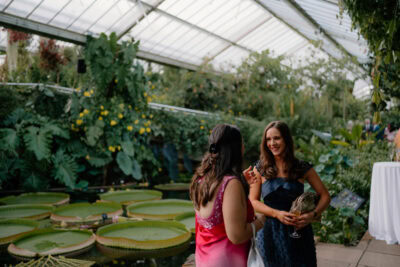 Women conversing in lush greenhouse setting.