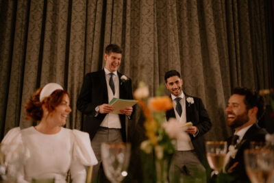 Wedding speech by groomsmen, guests smiling at reception.